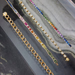Four bracelets displayed diagonally on marble: an 18K yellow gold chain, a gold bracelet with multicolored stones, a silver sapphire and diamond bracelet, and the Chroma Gradient Sapphire and Diamond Baguette Rainbow Tennis Bracelet in yellow gold.