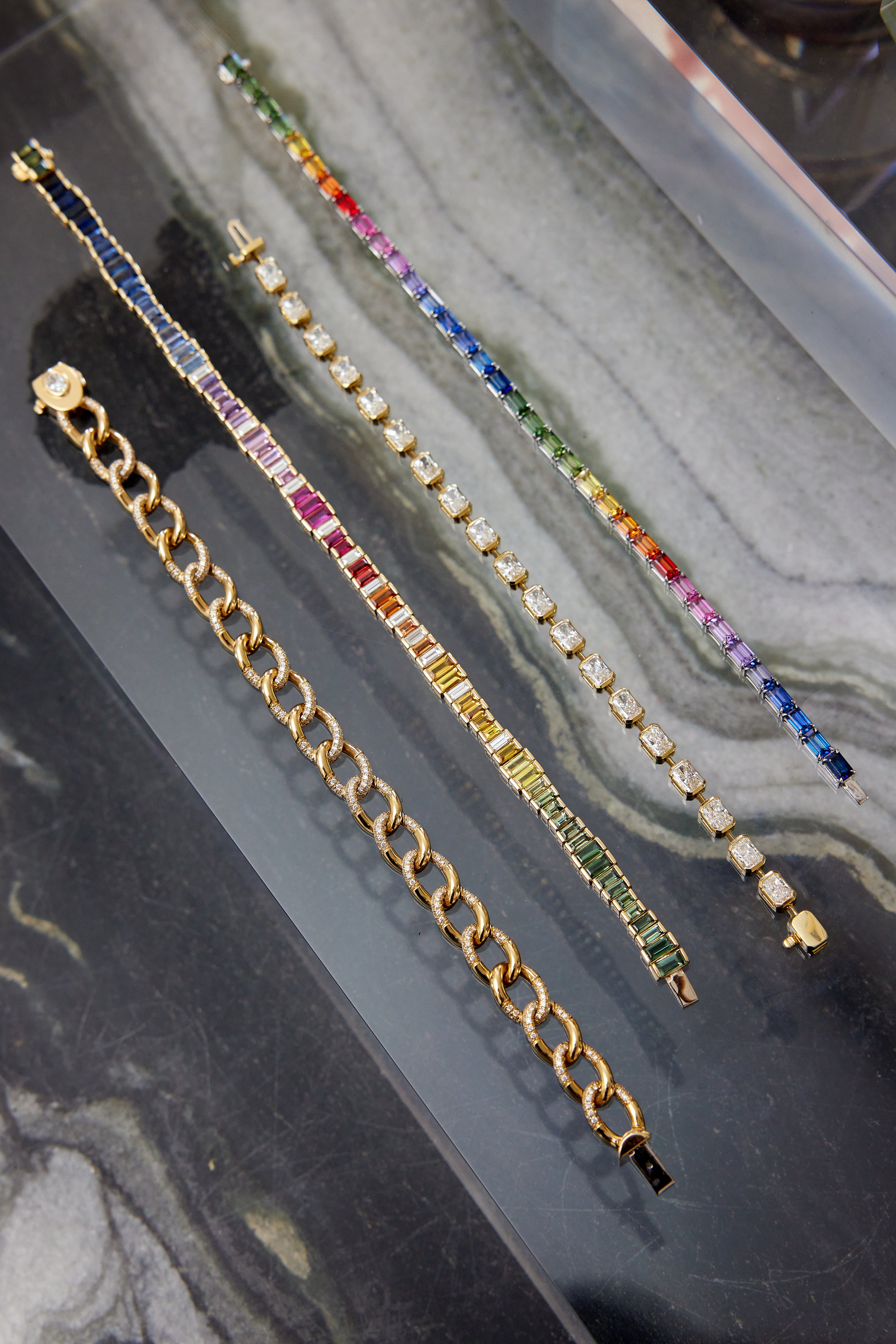 Four bracelets displayed diagonally on marble: an 18K yellow gold chain, a gold bracelet with multicolored stones, a silver sapphire and diamond bracelet, and the Chroma Gradient Sapphire and Diamond Baguette Rainbow Tennis Bracelet in yellow gold.