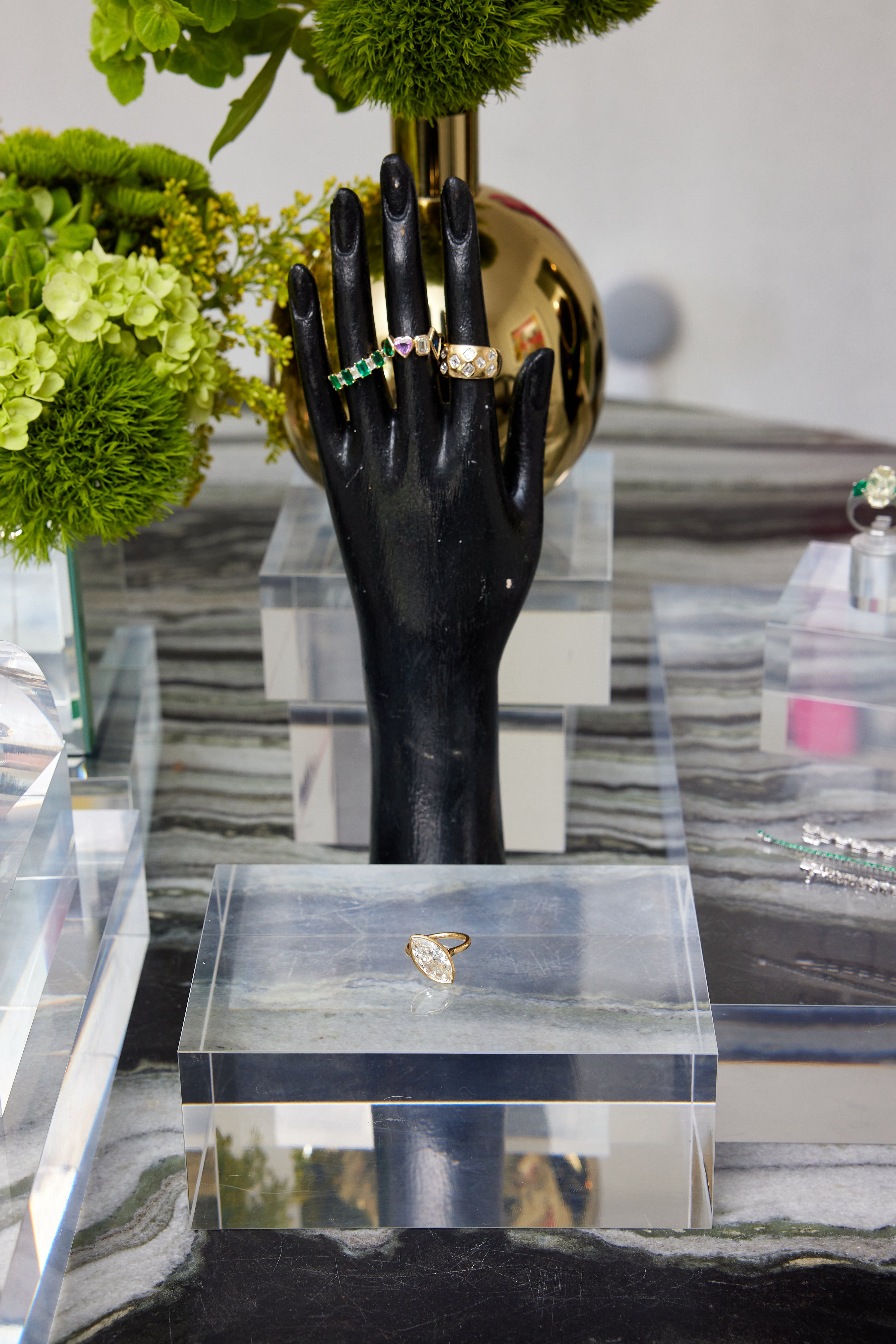 A black mannequin hand displays two 18K gold rings—one set with green gemstones and one with a large emerald cut diamond—among clear display blocks and green floral on stripes. Below, the Chroma Alternating Emerald and Diamond Half Eternity Band is displayed.
