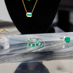 A display of emerald jewelry featuring the Chroma Alternating Emerald and Diamond Half Eternity Band, a ring, a necklace, and another ring with green gemstones, all arranged on marble with a black display stand in the background.