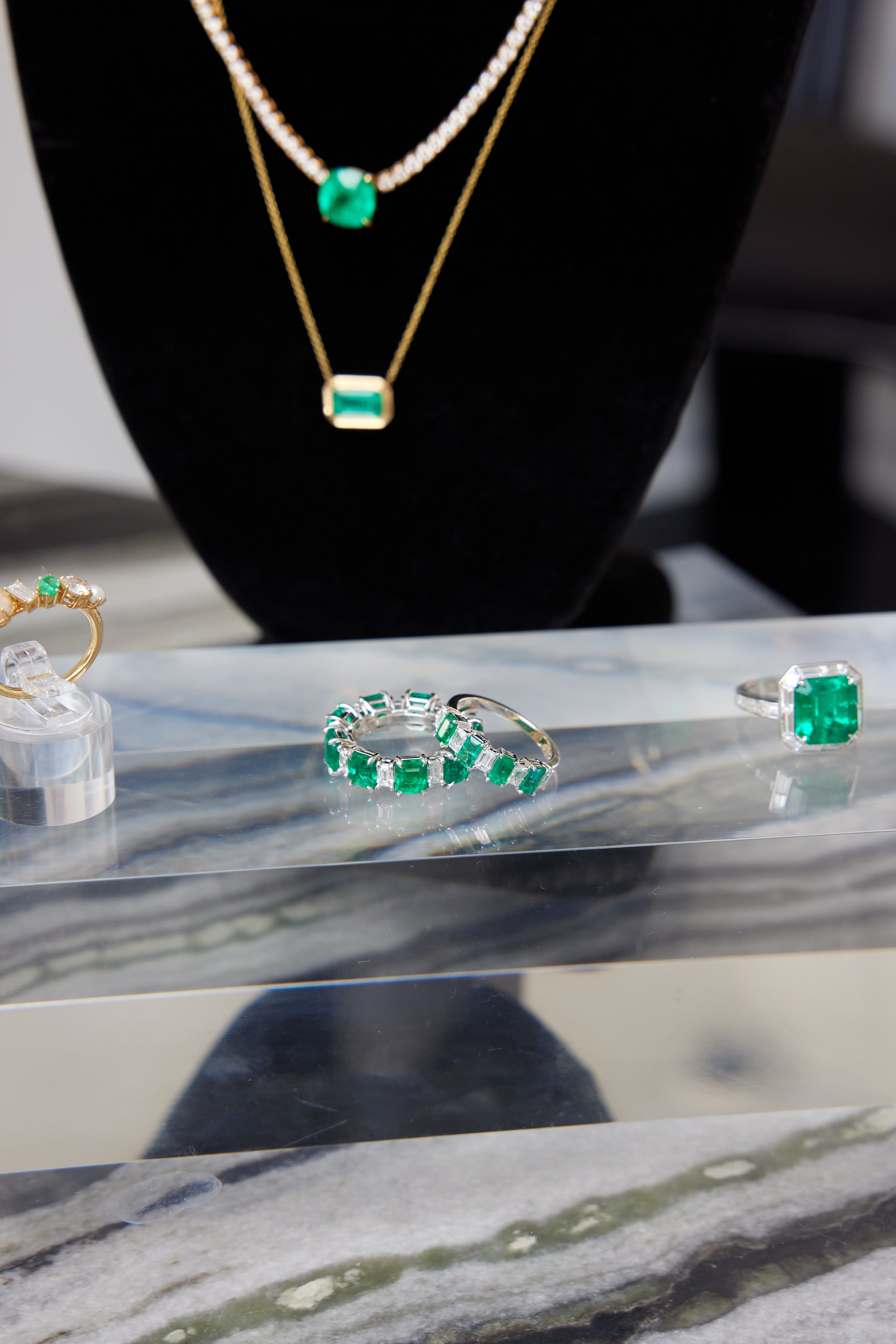A display of emerald jewelry featuring the Chroma Alternating Emerald and Diamond Half Eternity Band, a ring, a necklace, and another ring with green gemstones, all arranged on marble with a black display stand in the background.
