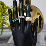 A black mannequin hand displays the Chroma Heart Beats Bezel Set Cluster Ring in yellow gold, featuring emerald, diamond, and sapphire, set against a blurred background with green plants and a reflective gold vase.