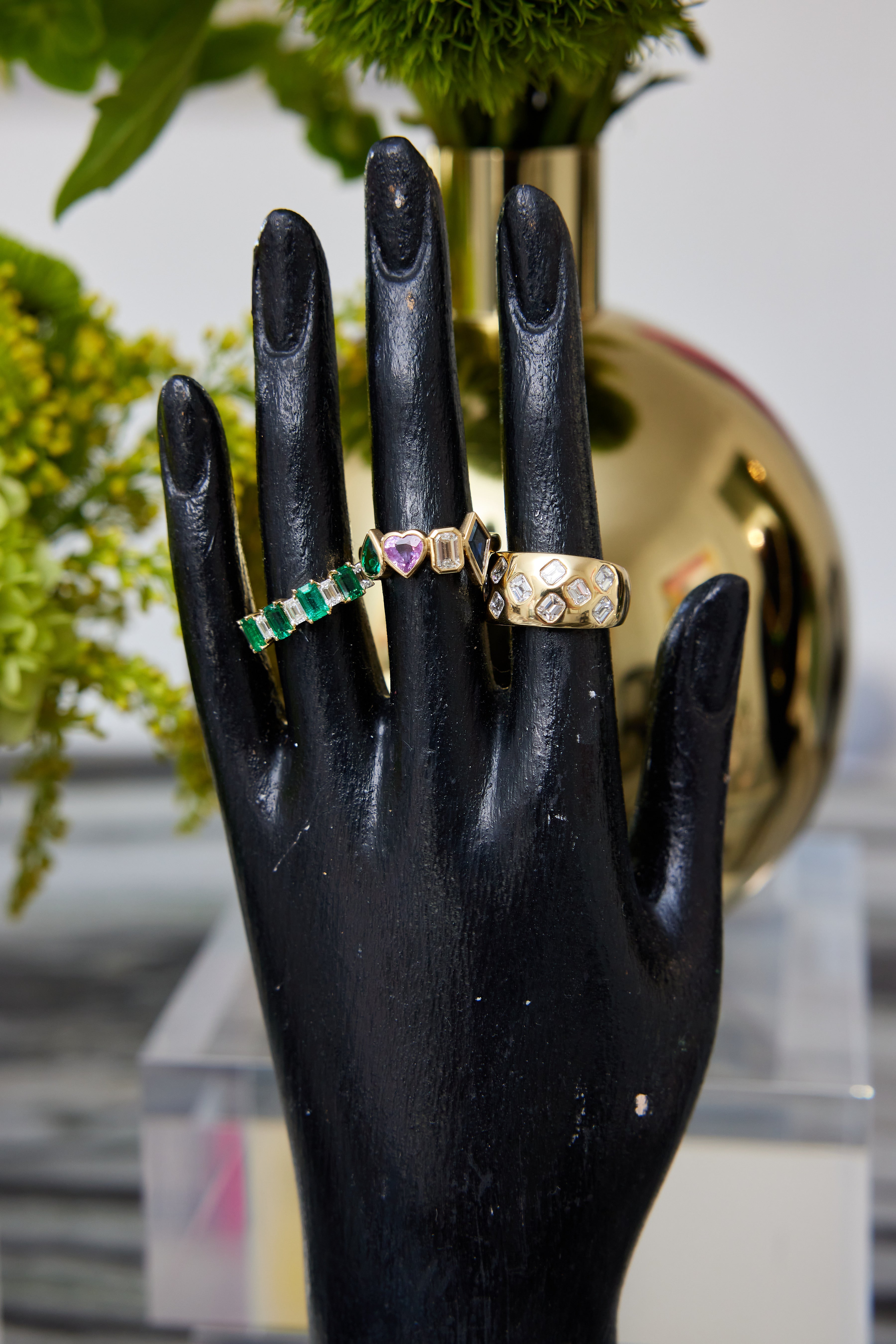 A black mannequin hand displays the Chroma Heart Beats Bezel Set Cluster Ring in yellow gold, featuring emerald, diamond, and sapphire, set against a blurred background with green plants and a reflective gold vase.