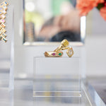The Chroma Stackable Dome Ring in yellow gold, featuring burnish set mixed shape sapphires and emeralds, is displayed on a clear acrylic stand with blurred florals and jewelry in the background.