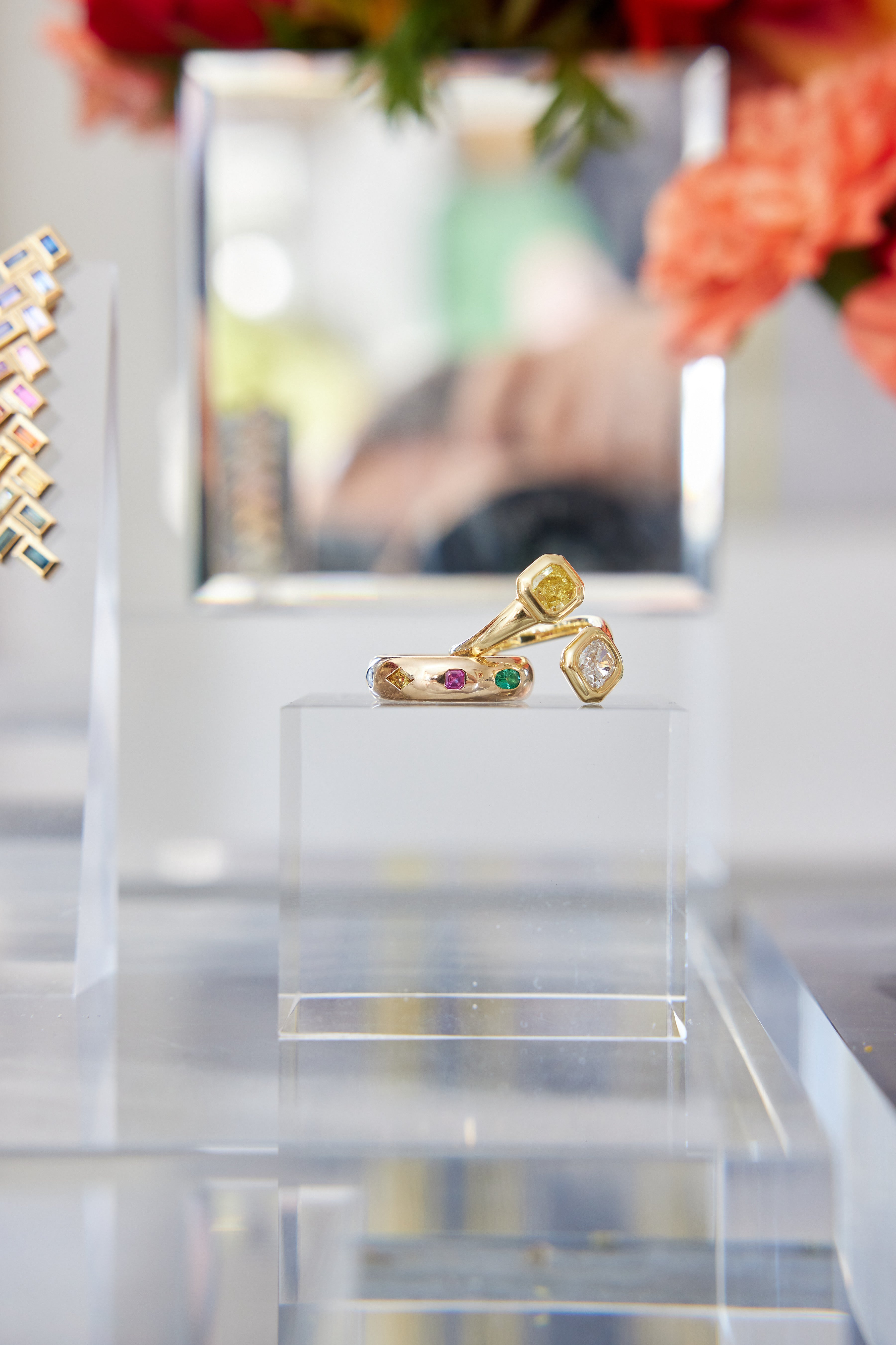 The Chroma Stackable Dome Ring in yellow gold, featuring burnish set mixed shape sapphires and emeralds, is displayed on a clear acrylic stand with blurred florals and jewelry in the background.