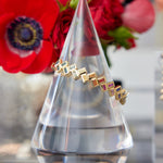 The Chroma Chevron Bezel Set Multicolor Baguette Sapphire Bangle in Yellow Gold is displayed on a clear, cone-shaped stand, with vibrant red flowers blurred in the background to highlight this elegant fine jewelry piece.