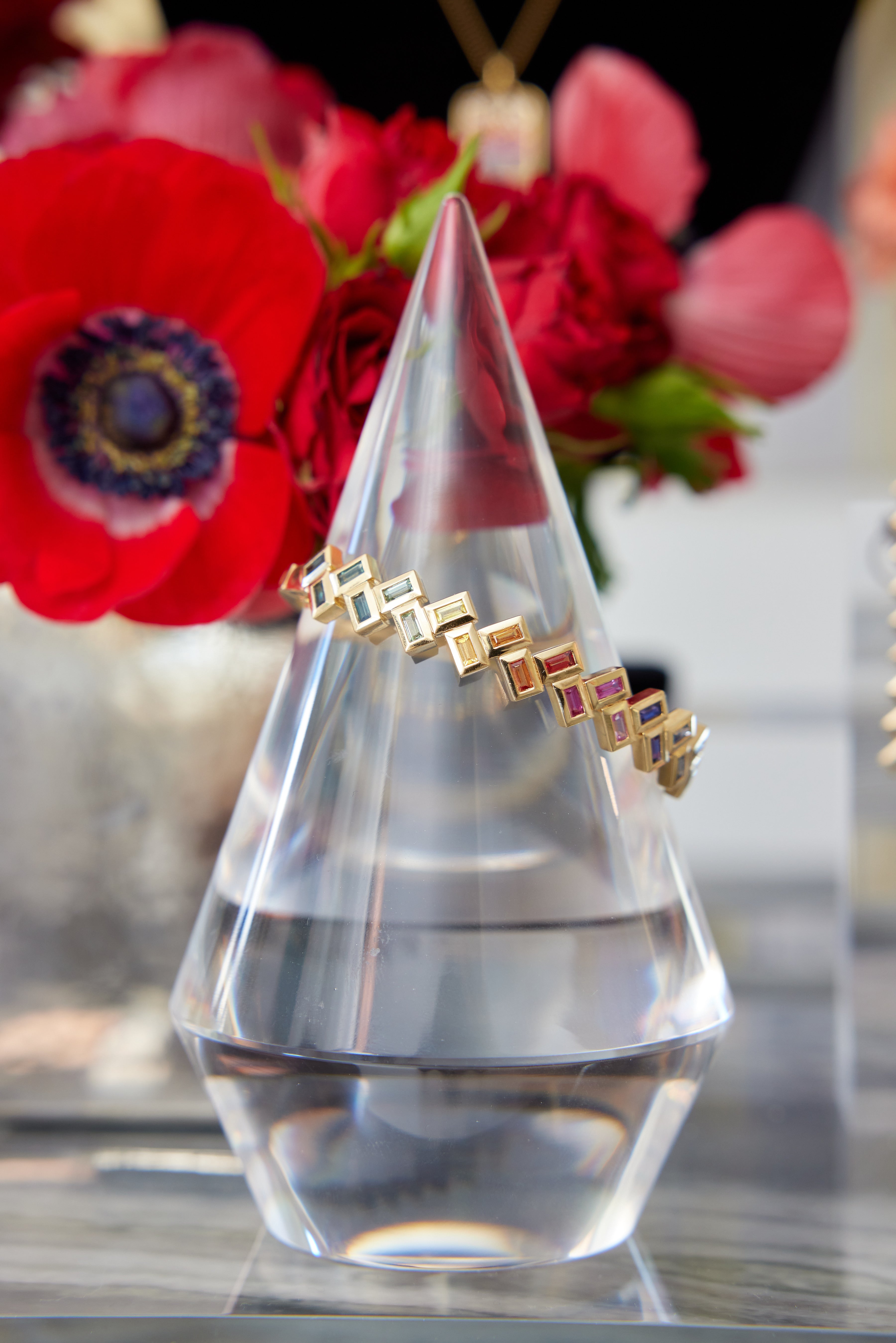 The Chroma Chevron Bezel Set Multicolor Baguette Sapphire Bangle in Yellow Gold is displayed on a clear, cone-shaped stand, with vibrant red flowers blurred in the background to highlight this elegant fine jewelry piece.