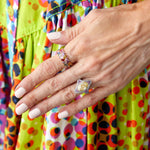 A close-up of a hand with pale pink manicured nails wears the Chroma Chevron Bezel Set Ring and colorful bands, resting on a vibrant dress with geometric circle patterns.