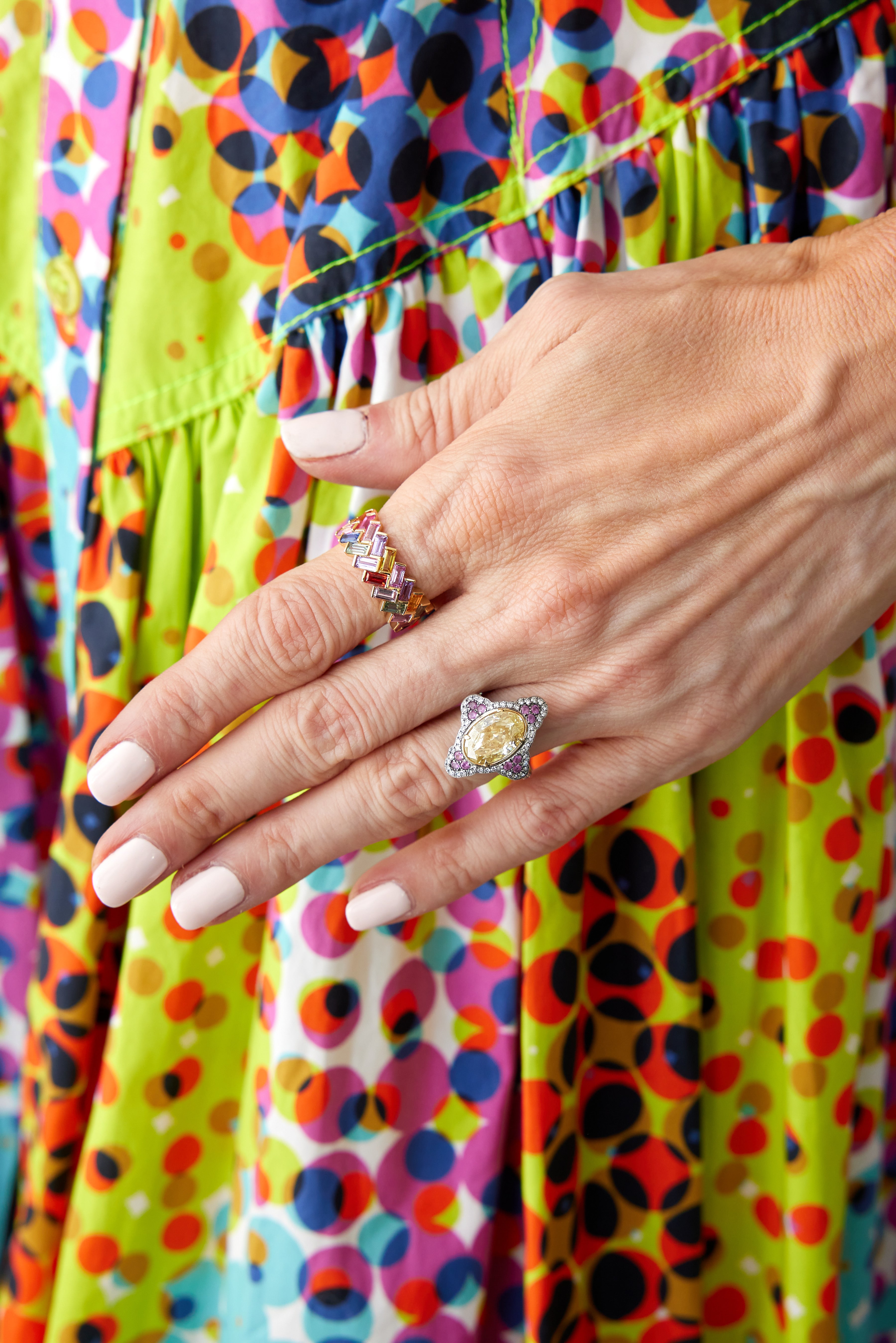 A close-up of a hand with pale pink manicured nails wears the Chroma Chevron Bezel Set Ring and colorful bands, resting on a vibrant dress with geometric circle patterns.