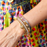 A wrist adorned with the Chroma Heart Beats Bezel Set Heart Shape Sapphire and Diamond Bangle in yellow gold among vibrant bracelets, with a multicolored polka dot skirt in the background.