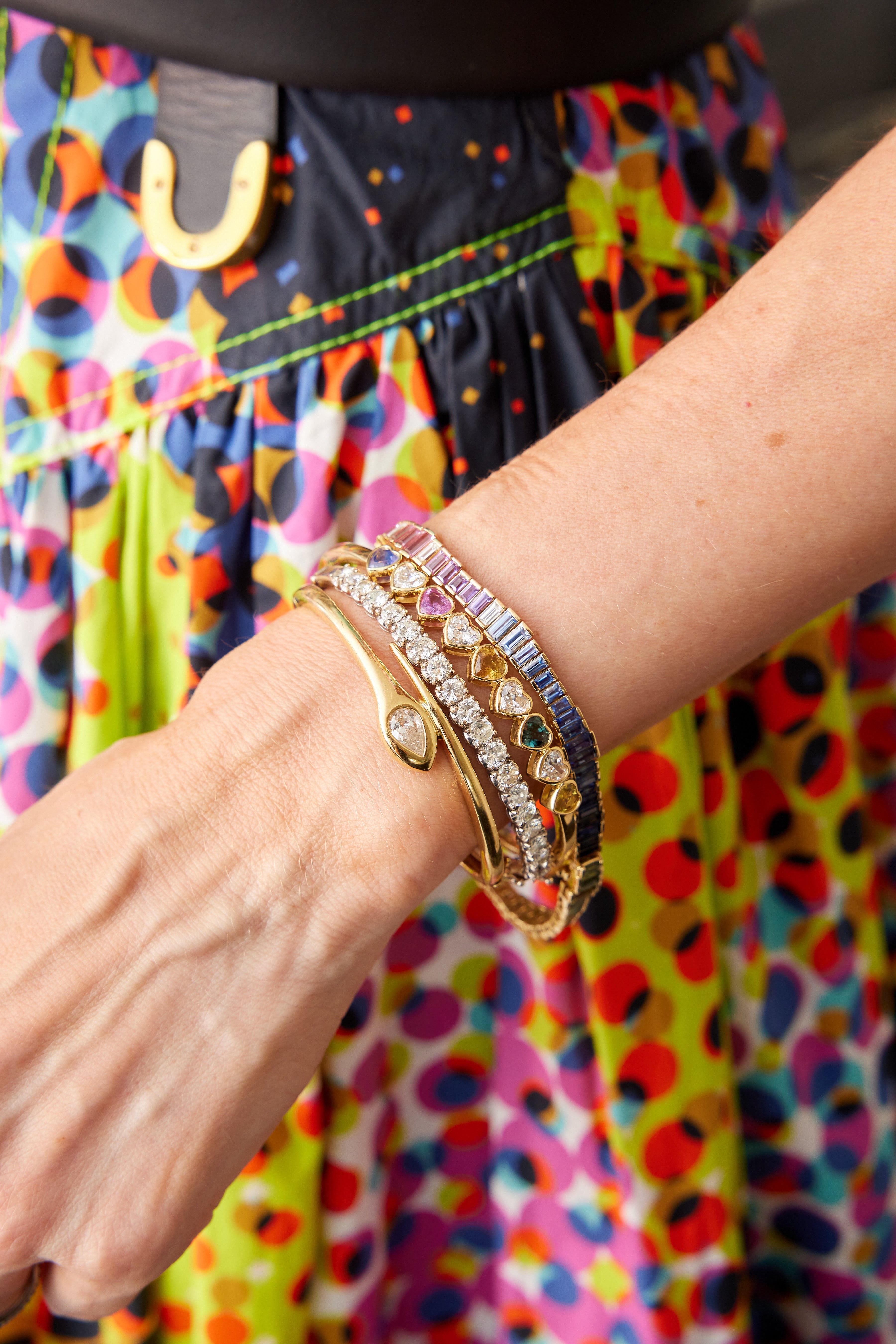A wrist adorned with the Chroma Heart Beats Bezel Set Heart Shape Sapphire and Diamond Bangle in yellow gold among vibrant bracelets, with a multicolored polka dot skirt in the background.