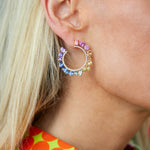 Close-up of a woman with blonde hair wearing Chroma Pavé Front-to-Back Hoops, colorful circular earrings with mixed shape sapphires set in 18K yellow gold. She wears a bright shirt with large orange dots and green details.