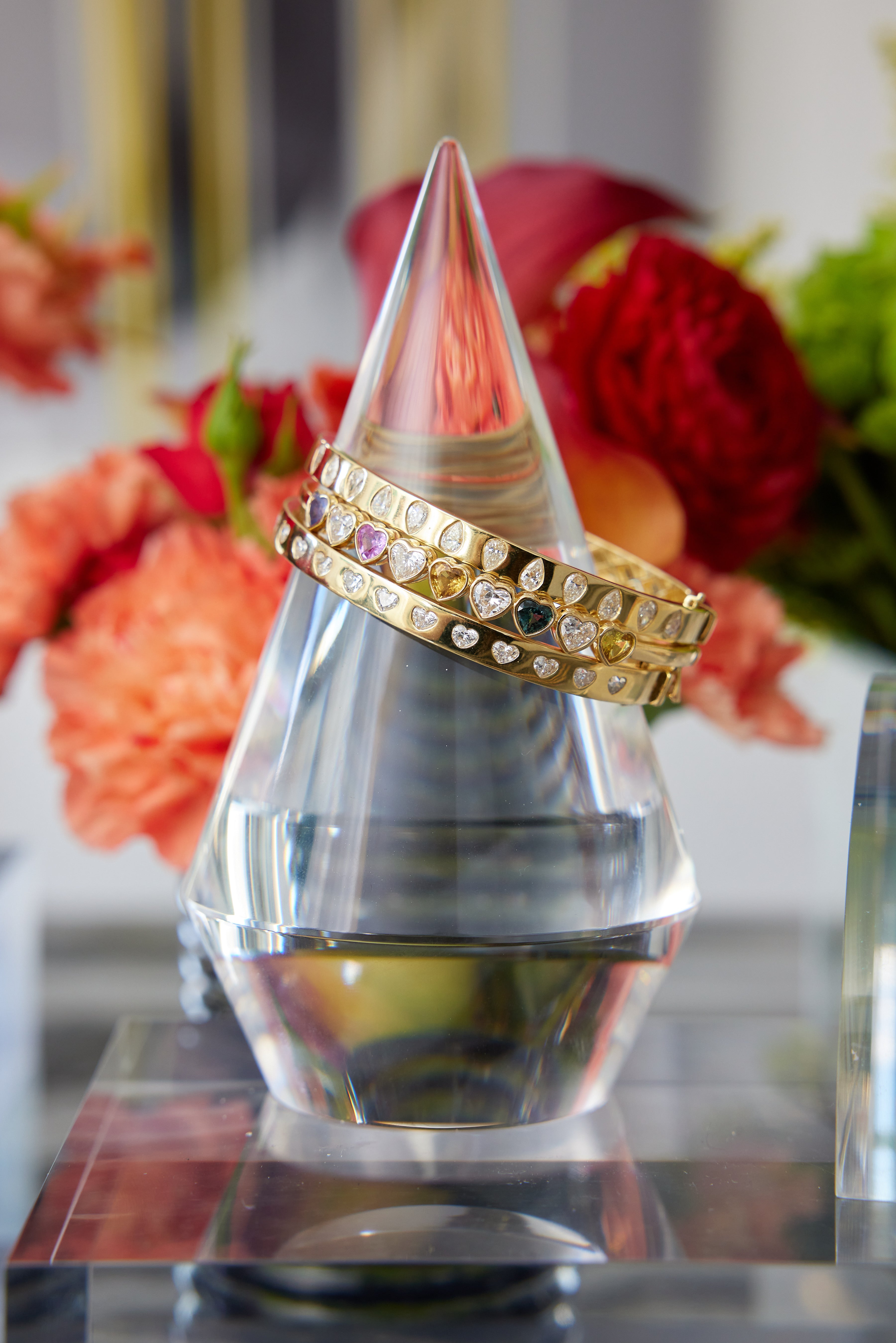 The Chroma Heart Beats Bezel Set Heart Shape Sapphire and Diamond Bangle in yellow gold rests on a clear, cone-shaped crystal with blurred red and peach flowers in the background.