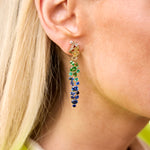 Close-up of a person with light blond hair wearing Multicolor Drop Earrings in yellow gold, featuring mixed shape sapphires and diamonds that sparkle in yellow, green, blue, and navy as the background softly blurs.