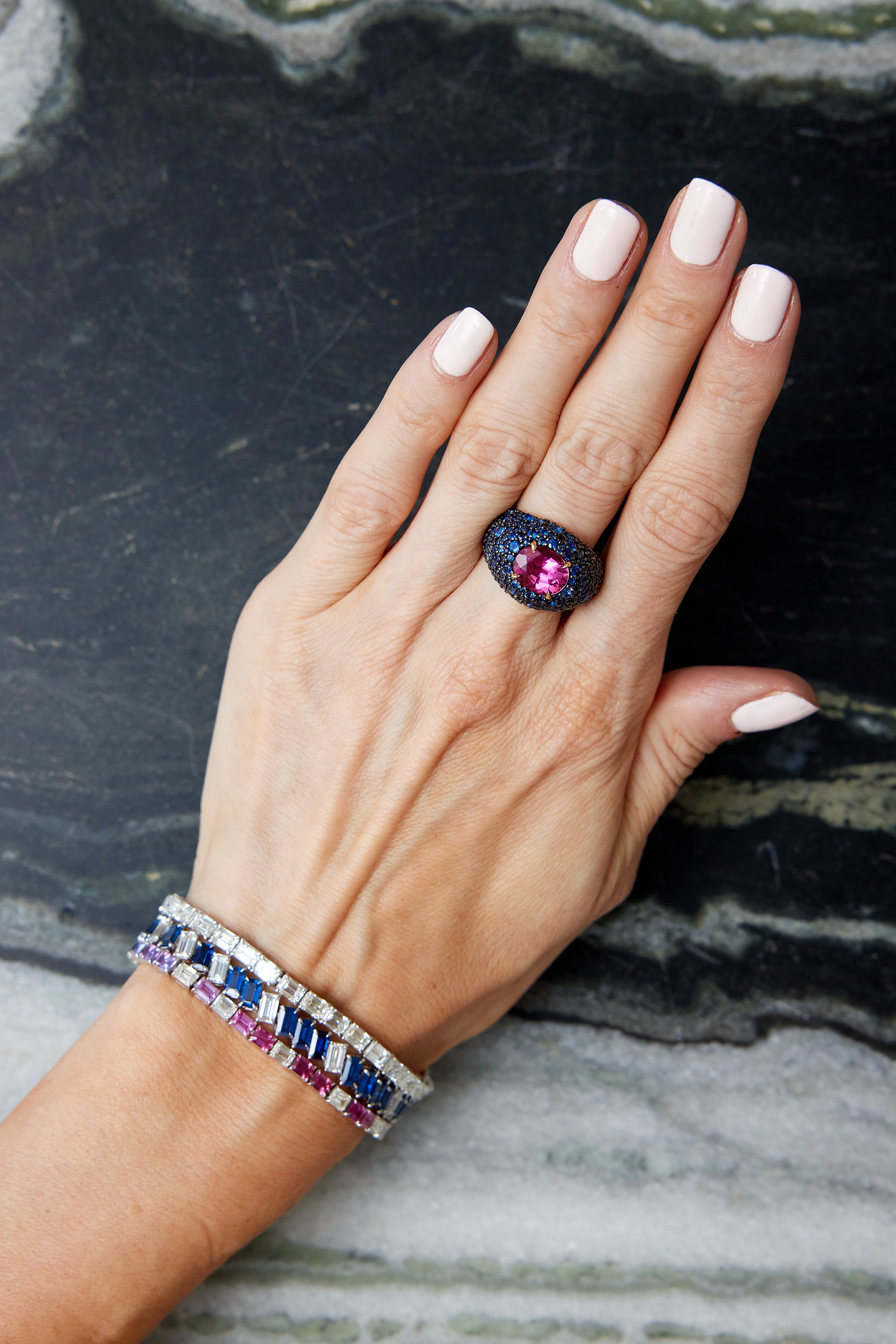 A hand with light pink nails rests on marble, showcasing the Chroma Empire Diagonally Set Emerald Cut Sapphire and Diamond Tennis Bracelet alongside a bold black ring with a large central pink stone.