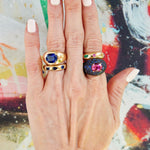 A hand with pale pink nail polish wears three chunky rings, including the Chroma Stackable Dome Ring in yellow gold with burnish set mixed shape sapphires and emeralds, posed against a vibrant, abstract background.