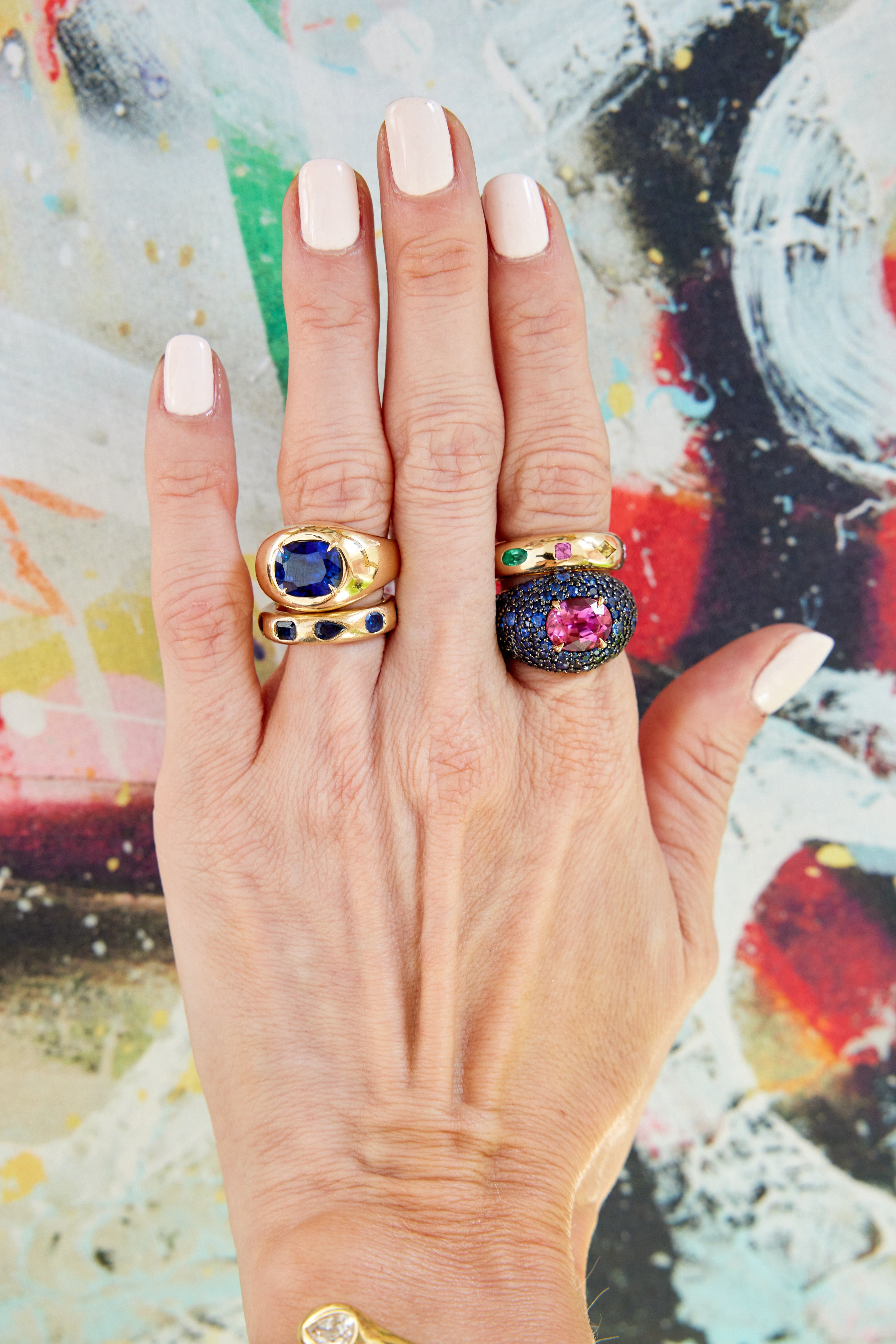A hand with pale pink nail polish wears three chunky rings, including the Chroma Stackable Dome Ring in yellow gold with burnish set mixed shape sapphires and emeralds, posed against a vibrant, abstract background.