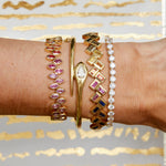 A forearm features four fine jewelry bracelets: an 18K yellow gold bangle with pink gemstones, a teardrop diamond bracelet, the Chroma Chevron Bezel Set Multicolor Baguette Sapphire Bangle in Yellow Gold, and one lined with clear diamonds. Gold and white background.