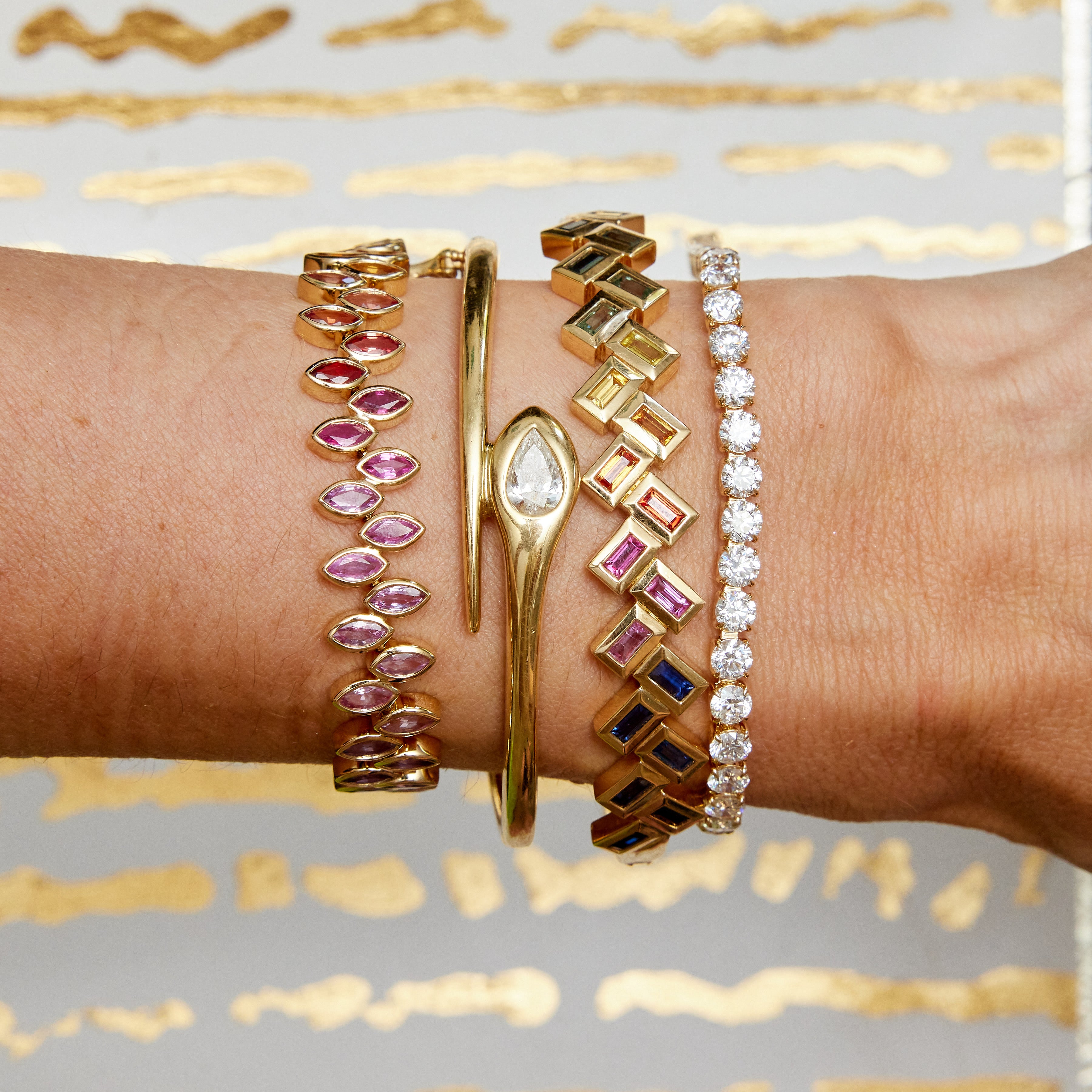 A forearm features four fine jewelry bracelets: an 18K yellow gold bangle with pink gemstones, a teardrop diamond bracelet, the Chroma Chevron Bezel Set Multicolor Baguette Sapphire Bangle in Yellow Gold, and one lined with clear diamonds. Gold and white background.