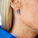 Close-up of a person with blonde hair and light skin wearing Chroma Empire Color Gradient Sapphire and Diamond Studs. The vibrant 18K white gold detail stands out against their bright blue collared shirt.