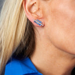 Close-up of a woman's ear with straight blonde hair, wearing Chroma Empire Color Gradient Sapphire and Diamond Studs. She is dressed in a bright blue collared shirt.