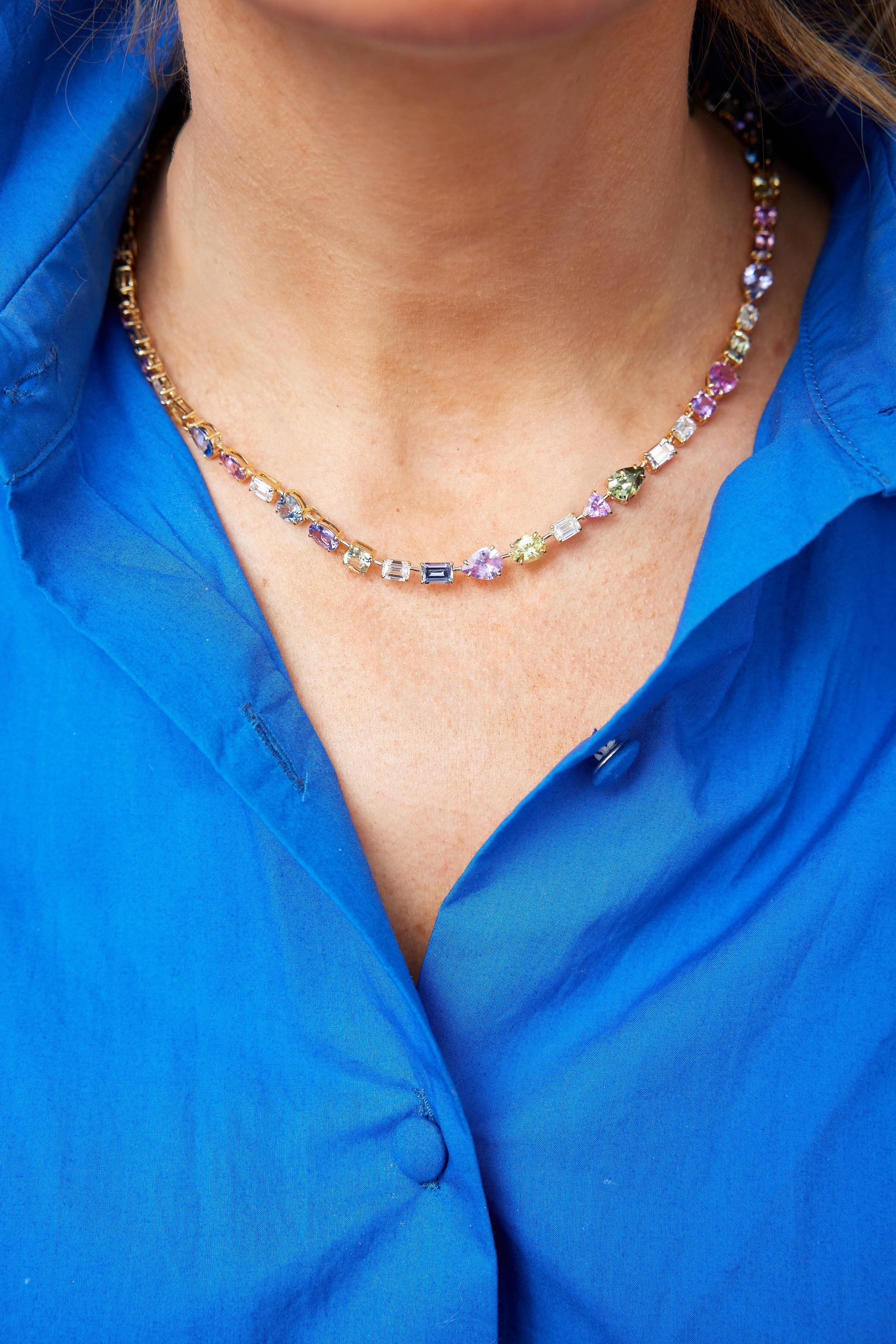 A person wearing a vibrant blue shirt and the Chroma Kaleidoscope Mixed Shape Sapphire and Diamond Tennis Necklace in Yellow Gold. The photo is cropped to show the neck, collar, and upper chest.