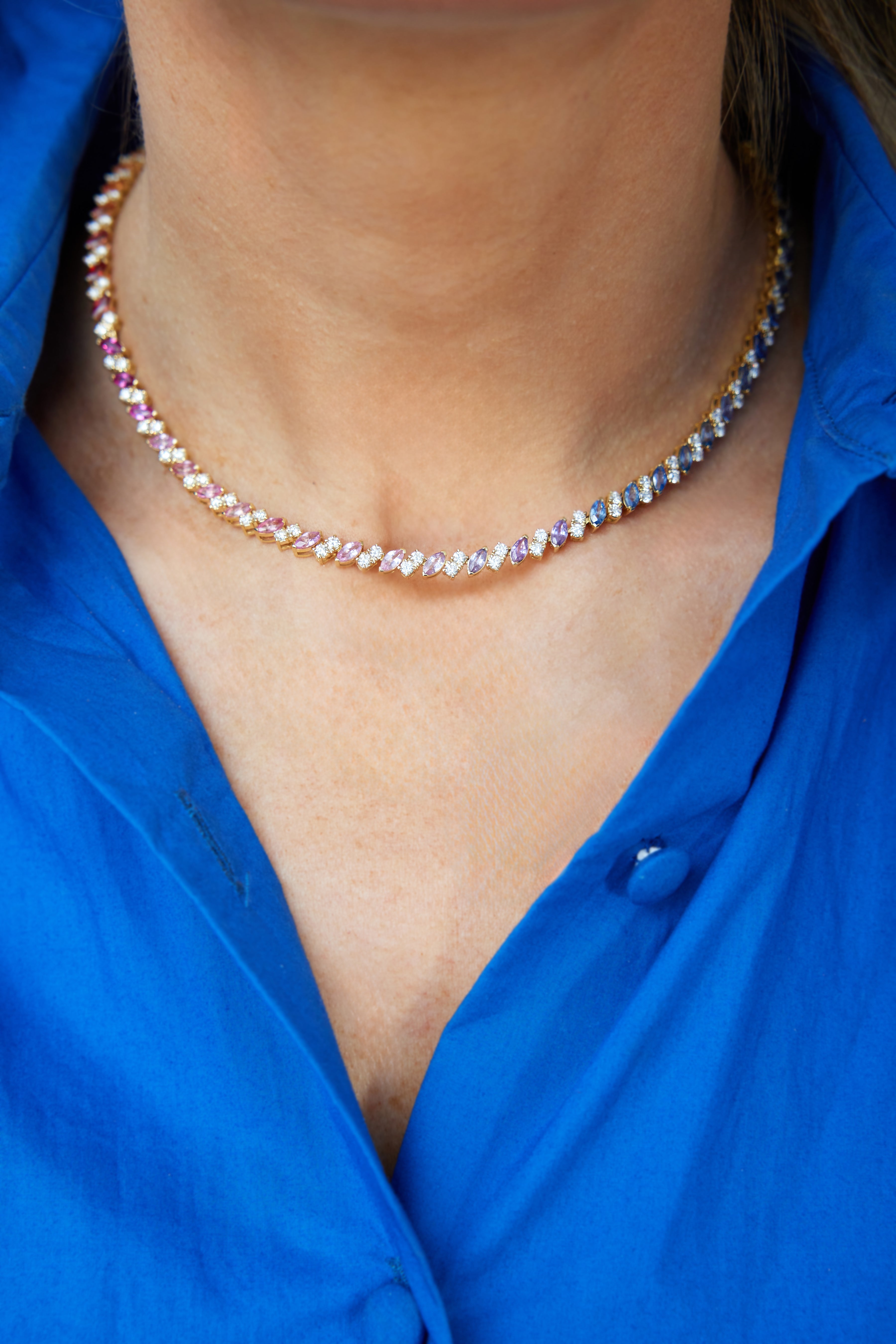 A close-up of a person wearing the Chroma Match Point Multicolor Tennis Necklace in yellow gold with marquise sapphires and round diamonds, paired with a vibrant blue button-up shirt—an eye-catching fine jewelry piece.