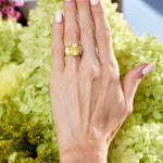 A hand with pale pink manicured nails rests on light green hydrangeas, wearing a large yellow gemstone ring and the Chroma Kaleidoscope Mixed Shape Sapphire Tennis Bracelet in Yellow Gold adorned with vibrant sapphires.