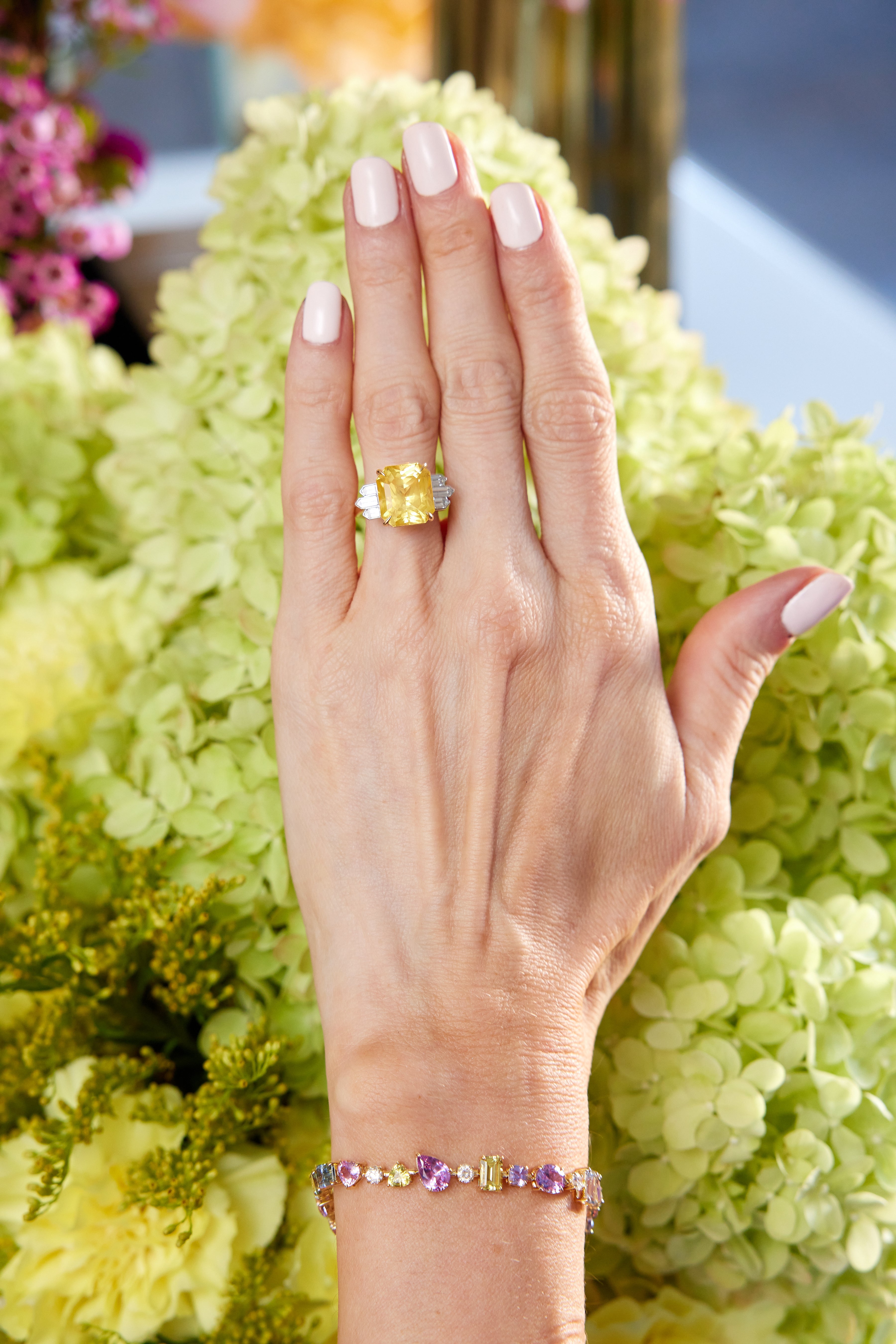 A hand with pale pink manicured nails rests on light green hydrangeas, wearing a large yellow gemstone ring and the Chroma Kaleidoscope Mixed Shape Sapphire Tennis Bracelet in Yellow Gold adorned with vibrant sapphires.