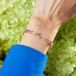 A wrist adorned with the Chroma Kaleidoscope Mixed Shape Sapphire Tennis Bracelet in Yellow Gold and a vivid blue sleeve, set against pale green flowers—a vibrant showcase of maximalist style.