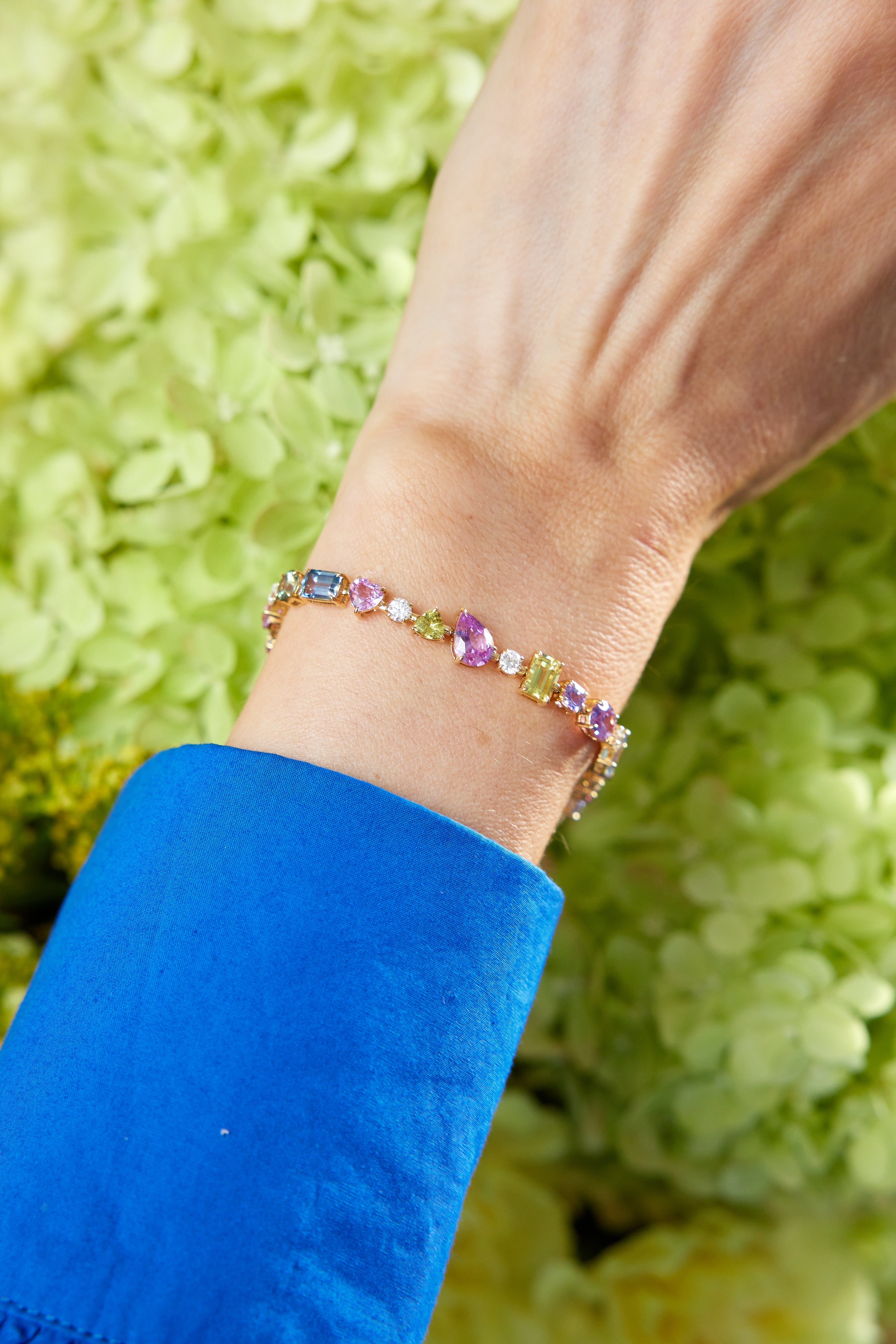 A wrist adorned with the Chroma Kaleidoscope Mixed Shape Sapphire Tennis Bracelet in Yellow Gold and a vivid blue sleeve, set against pale green flowers—a vibrant showcase of maximalist style.