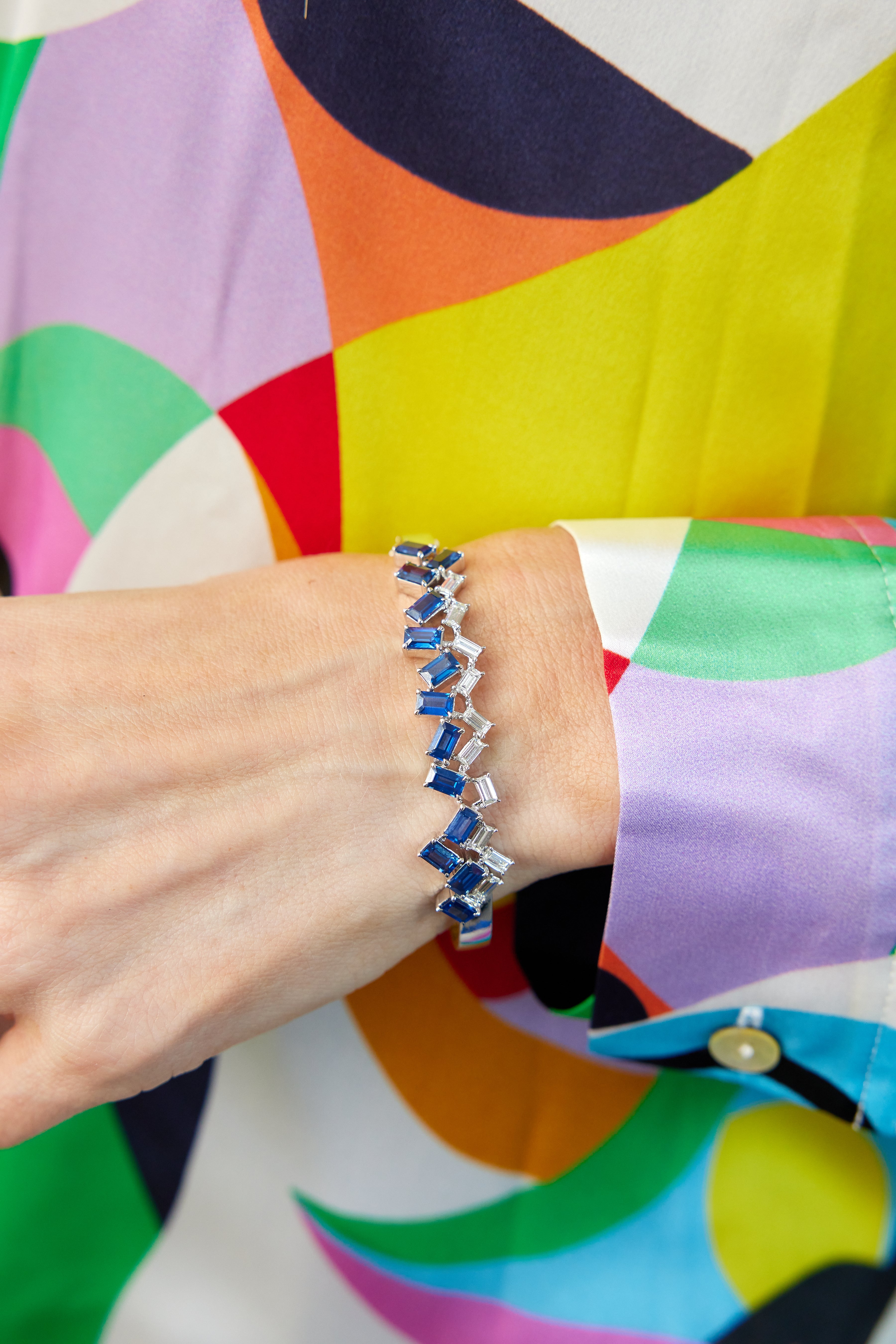 A person wearing a colorful, patterned shirt with a striking Chroma Empire Alternating Emerald Cut Sapphire and Diamond Bangle in White Gold on their wrist.