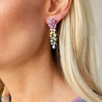Close-up of a woman with blonde hair wearing Chroma Kaleidoscope Drop Earrings in yellow gold, featuring mixed shape multicolor sapphires and diamonds. She pairs them with a vibrant, patterned top.