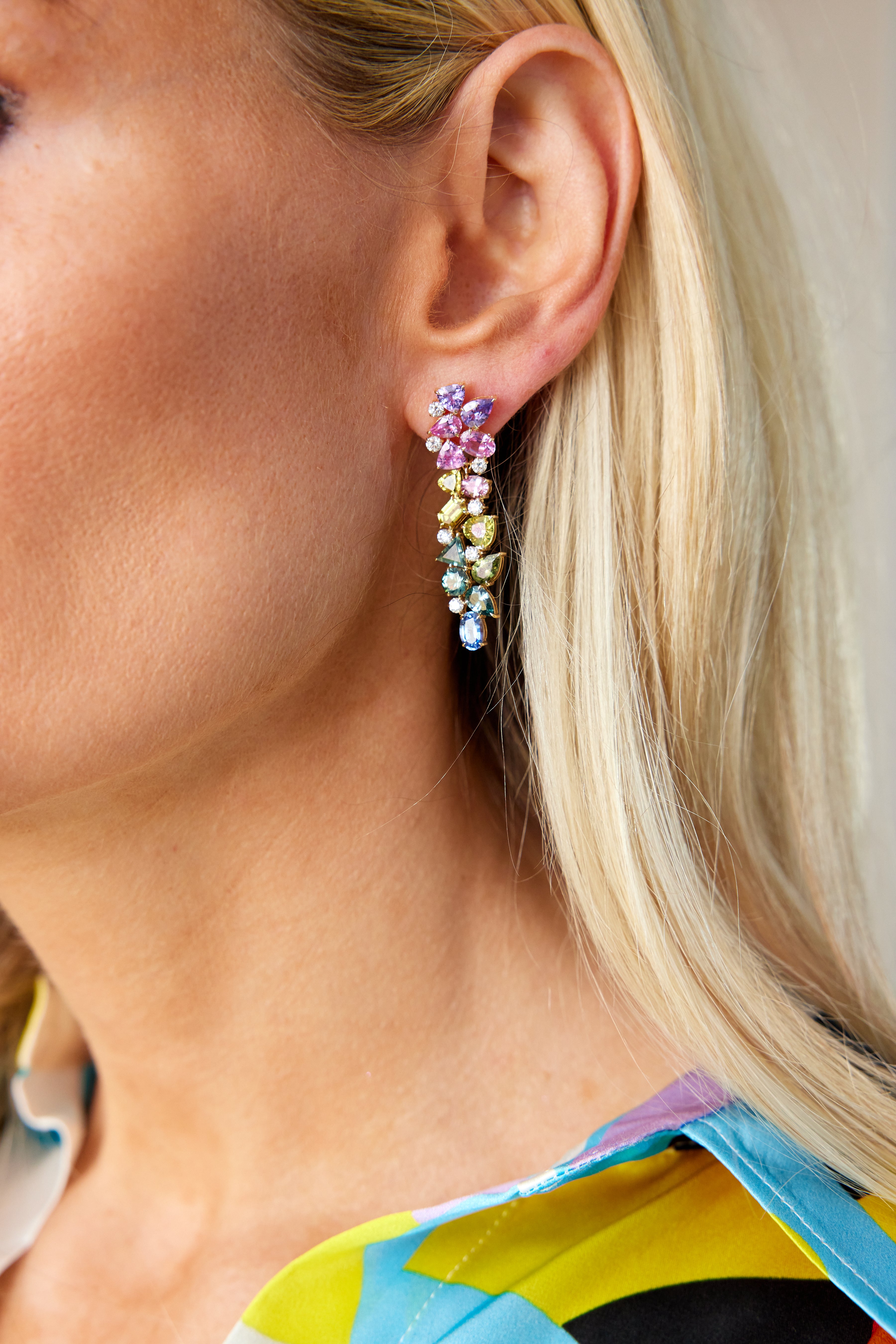 Close-up of a woman with blonde hair wearing Chroma Kaleidoscope Drop Earrings in yellow gold, featuring mixed shape multicolor sapphires and diamonds. She pairs them with a vibrant, patterned top.