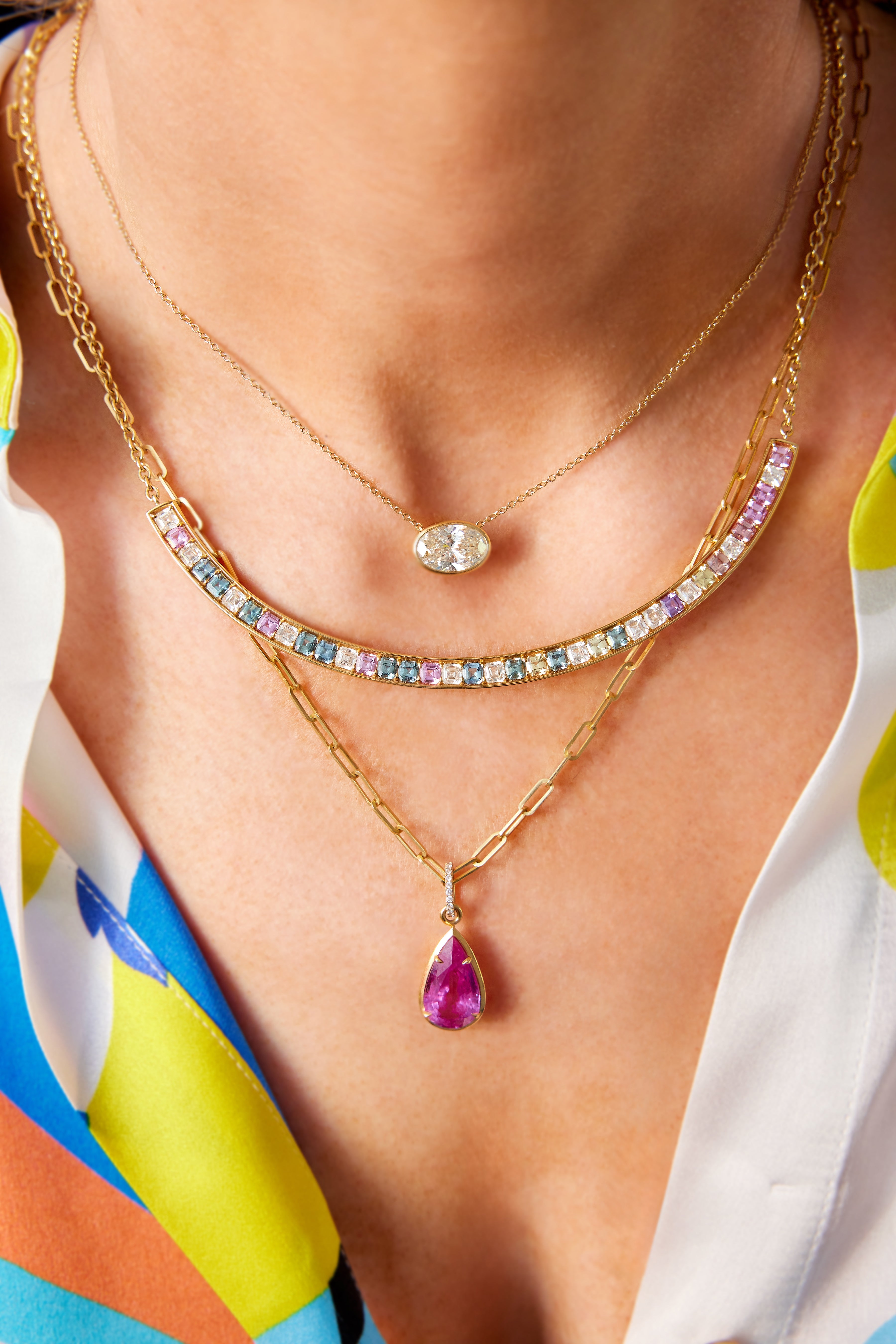 A close-up of a person wearing the Ombré Smile Bar Necklace in yellow gold with Asscher cut diamonds and multicolor sapphires, styled with layered gold chains and a colorful patterned shirt.