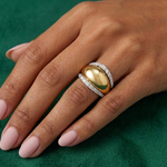 A hand with light pink manicured nails wears the Coil Dome Ring with Pavé Diamond Borders in Yellow Gold, featuring a bold dome design flanked by diamond rows, shown on a green textured surface.