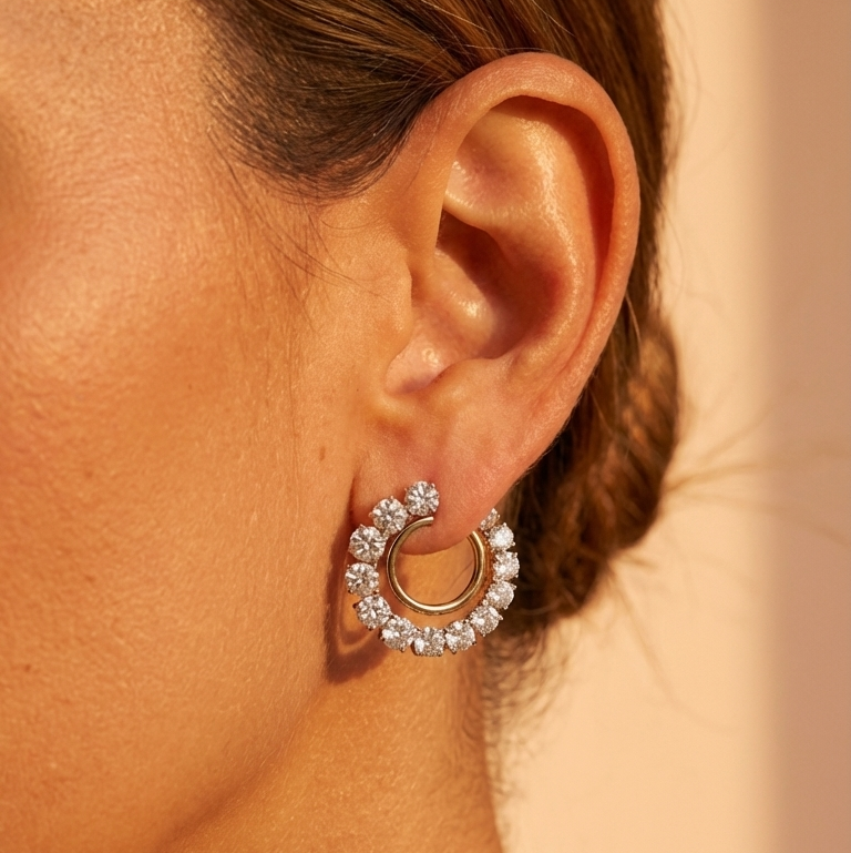 A close-up of a woman's ear wearing Coil Round Diamond Front-to-Back Hoop Earrings in yellow gold and platinum. Sparkling diamonds catch the light against a warmly lit background, showcasing the elegance of these stylish hoops.