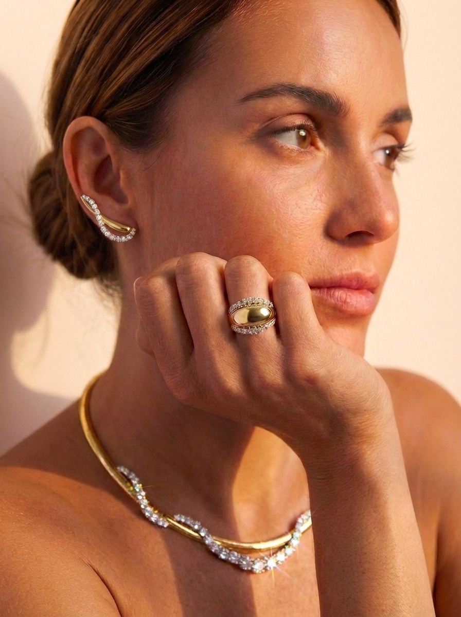 A woman with brown hair in a low bun wears the Coil Graduated Round Diamond and Omega Chain Necklace in Yellow Gold and Platinum, along with a distinctive ring and curved diamond earring, her hand resting on her chin as she gazes thoughtfully.