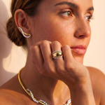 A woman with brown hair in a low bun wears the Coil Graduated Round Diamond and Omega Chain Necklace in Yellow Gold and Platinum, along with a distinctive ring and curved diamond earring, her hand resting on her chin as she gazes thoughtfully.