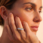 A woman with her hair tied back gazes thoughtfully, resting her chin on her hand and showcasing the Coil Graduated Round Diamond Wave Band in Yellow Gold, adorned with sparkling natural diamonds.