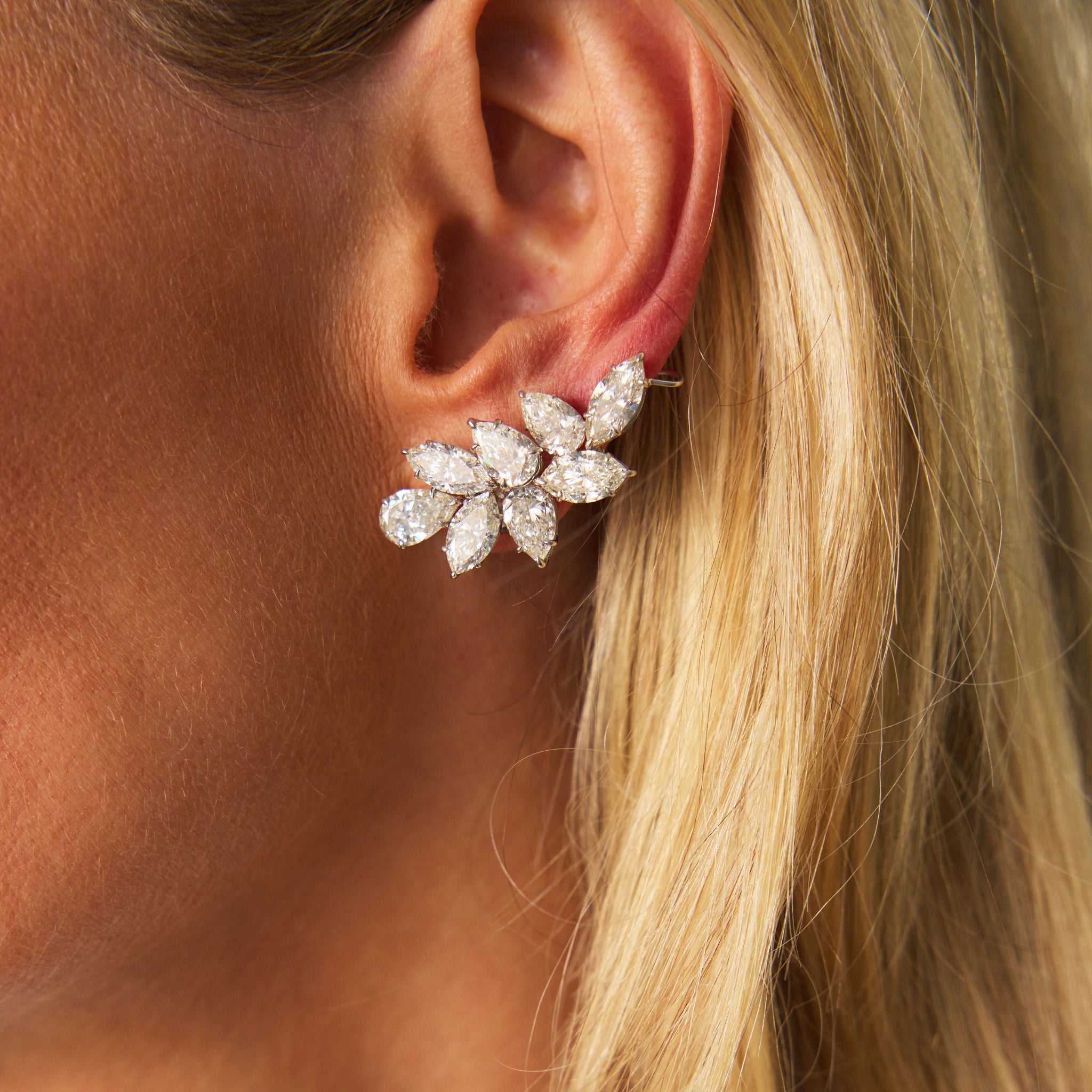 Close-up of a woman's ear wearing a Pair of Diamond Earclips crafted in 18K white gold, featuring large floral-shaped clusters with marquise-cut diamonds. Her straight blonde hair is tucked behind her ear.