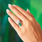 A hand with manicured nails wears the Early 20th Century Emerald And Diamond Bypass Ring, featuring a teardrop Colombian emerald and a diamond-adorned curved silver band, set against a blurred green background.