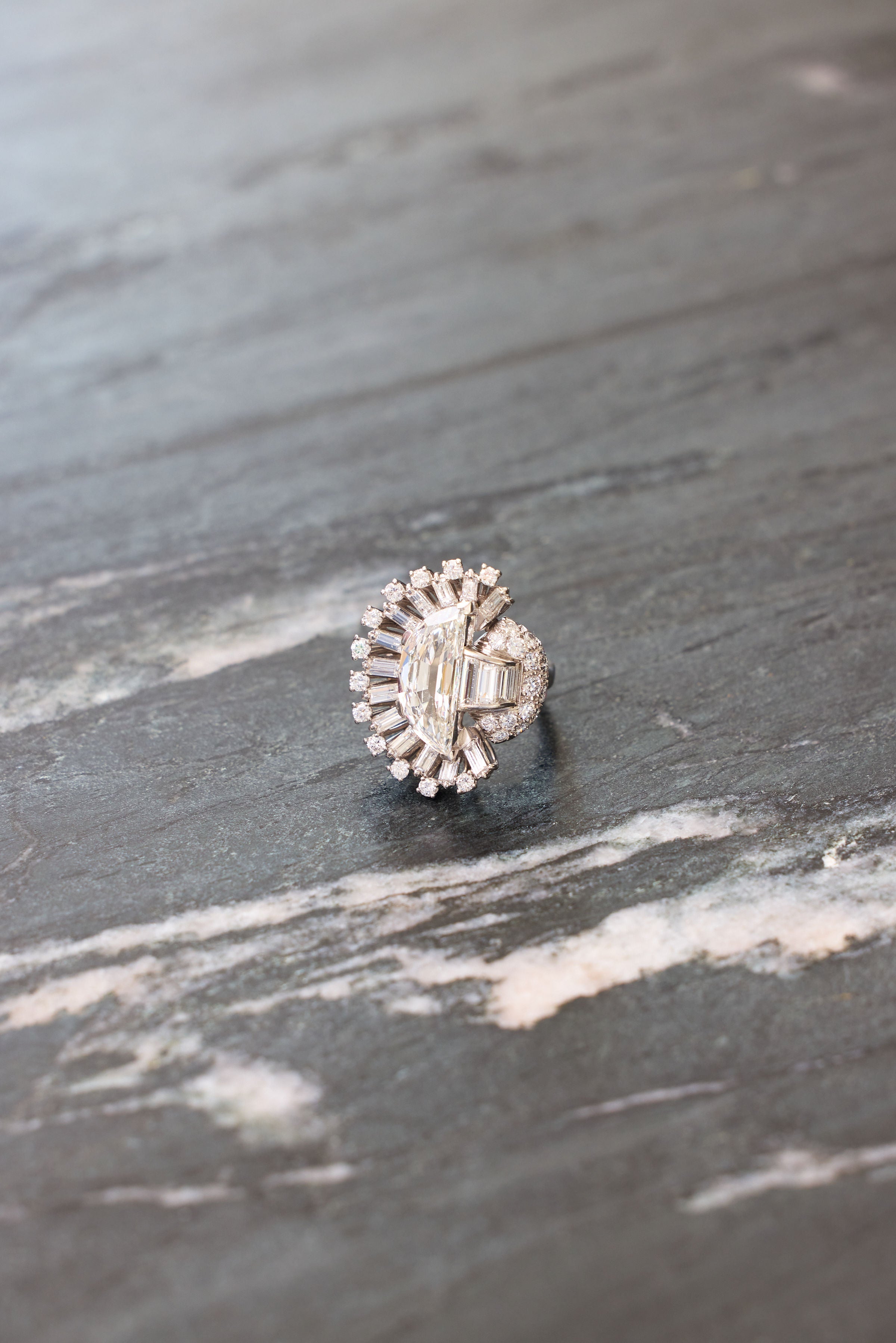 The Oscar Heyman & Brothers Diamond Ring, crafted in 1946 with baguette, round, and half-moon diamonds in platinum and a unique fan-shaped design, is displayed on a gray marble surface.