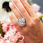 A hand with light pink manicured nails wears the Vintage Oscar Heyman & Brothers Diamond Ring, an ornate platinum piece from 1946, featuring clear gemstones and half-moon diamonds in the classic Oscar Heyman style, posed near white and pink flowers.