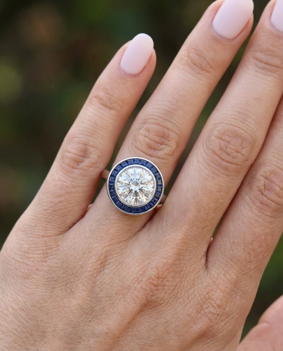 A hand with neatly manicured, pale pink nails wears the Round Brilliant Diamond with Channel Set Sapphire Halo Ring, featuring a round central diamond and a sapphire halo on a white gold or silver band.