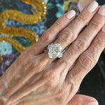A hand with light pink nails wears the Oval Diamond Engagement Ring with Heart Shape Side Stones, featuring a large oval diamond and two heart-shaped side stones on a platinum band. A gold textured object is blurred in the background.