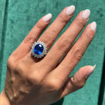 A hand with light pink nails showcases the Sapphire and Diamond Ring, featuring a large cushion-cut sapphire surrounded by small clear stones in a silver setting, against a green textured background.