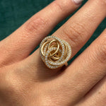 A close-up of a hand wearing the Gold and Diamond Ring, featuring an intricate interwoven gold design with sparkling diamonds along the edges, set against a deep green background.