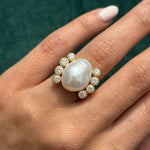 A hand wears the Cultured Pearl and Diamond Ring, featuring a large irregular white pearl center surrounded by sparkling round diamonds, set against a dark green background.
