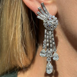 Close-up of a woman's ear wearing the Pair of Diamond Earclips, featuring large, intricate platinum earrings with round, baguette, and teardrop diamonds in a cascading 1950s-inspired design. Blonde hair is partially visible in the background.